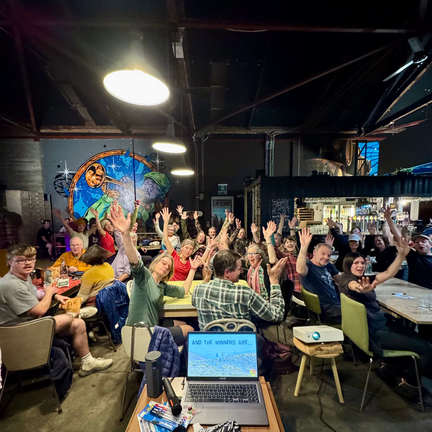 A large group of people sitting at tables in a dimly lit loft-style room, raising their hands enthusiastically. In the foreground, a wooden table holds a laptop, notebooks, and other items.